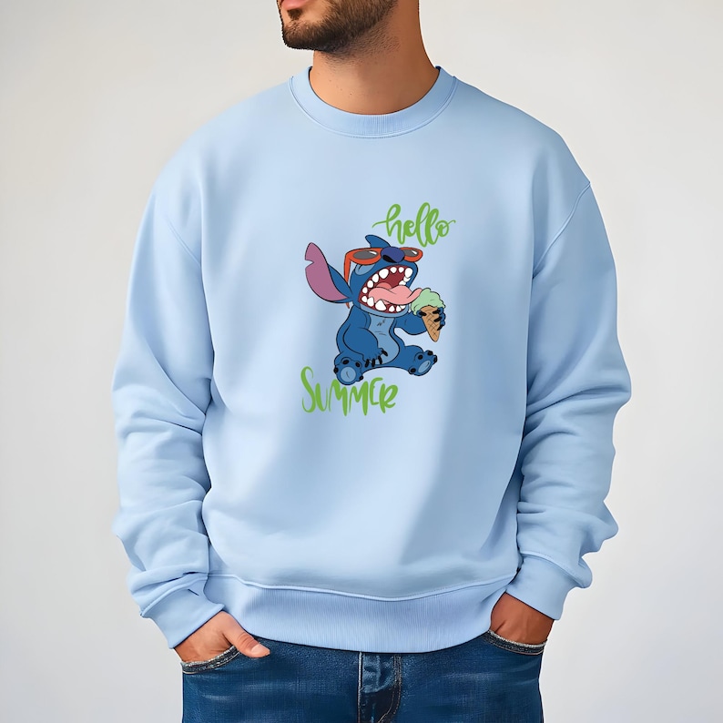Puede incluir: Sudadera azul claro con un personaje de dibujos animados, Stitch, con gafas de sol y un helado. Las palabras "hello Summer" est&aacute;n escritas en verde arriba y abajo del personaje. La sudadera es de color azul claro liso.