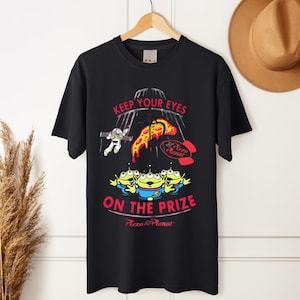 May include: Black t-shirt featuring a graphic design with the text "KEEP YOUR EYES ON THE PRIZE" and "Pizza Planet" logo. The design includes cartoon aliens, Buzz Lightyear, and pizza slices.