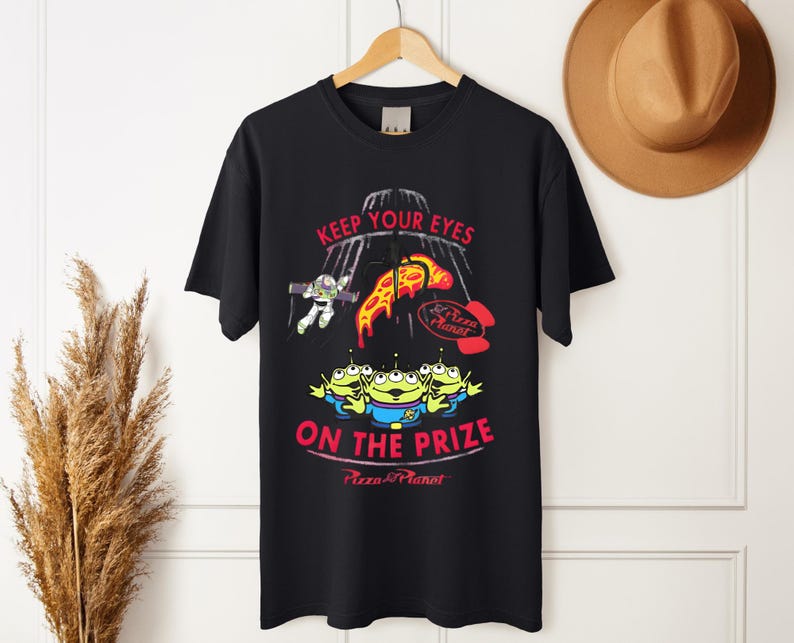 May include: Black t-shirt featuring a graphic design with the text "KEEP YOUR EYES ON THE PRIZE" and "Pizza Planet" logo. The design includes cartoon aliens, Buzz Lightyear, and pizza slices.