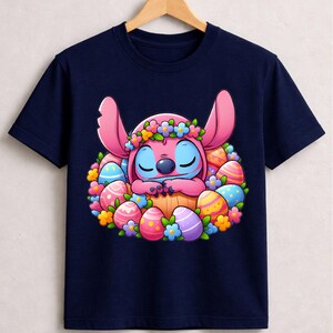 May include: Navy blue t-shirt featuring a cartoon character with pink ears and blue eyes, surrounded by colorful Easter eggs and flowers. The character is wearing a flower crown. The design is centered on the shirt.