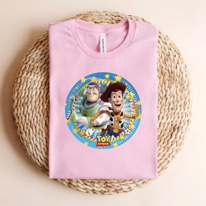 Puede incluir: Una camiseta rosa claro con un gr&aacute;fico circular de Buzz Lightyear y Woody de Toy Story. El gr&aacute;fico est&aacute; rodeado por un borde azul con estrellas amarillas y las palabras "Toy Story". La camiseta est&aacute; doblada sobre una superficie tejida.