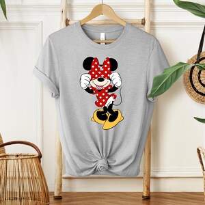 Pu&ograve; includere: Una maglietta grigia con una grafica di Minnie Mouse. Minnie &egrave; raffigurata nel suo outfit classico: un vestito rosso a pois bianchi, un fiocco abbinato e scarpe gialle. La maglietta &egrave; annodata in vita e appesa a una gruccia di legno.