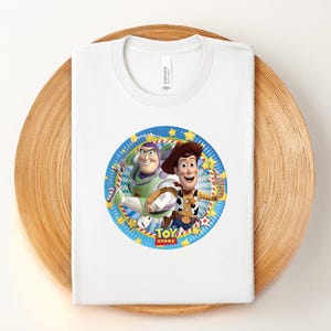 Puede incluir: Camiseta blanca con un dise&ntilde;o circular de Buzz Lightyear y Woody de Toy Story. El dise&ntilde;o tiene un borde azul y amarillo y las palabras "Toy Story". La camiseta est&aacute; doblada sobre una superficie de madera.