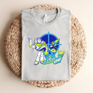 May include: A pale grey t-shirt with a vibrant graphic of Buzz Lightyear and the Little Green Men from Toy Story. The design includes a blue target, a rocket ship, and the words "Star Command".