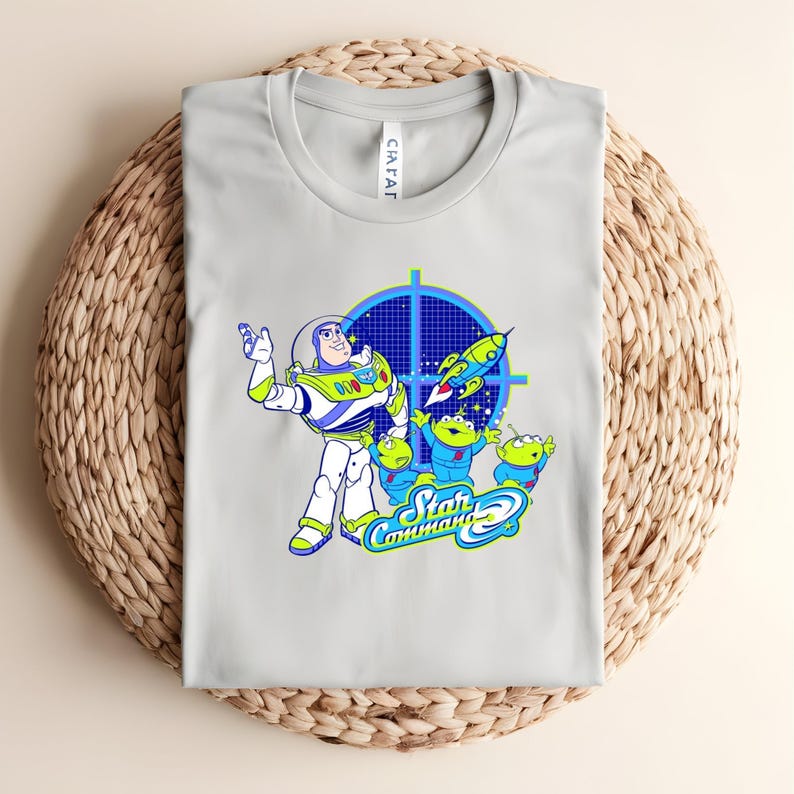 May include: A pale grey t-shirt with a vibrant graphic of Buzz Lightyear and the Little Green Men from Toy Story. The design includes a blue target, a rocket ship, and the words "Star Command".