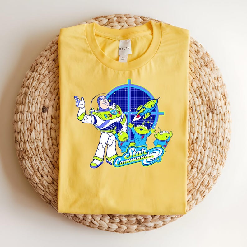 May include: Yellow t-shirt with a graphic of Buzz Lightyear and the Little Green Men from Toy Story. The design includes a blue target, a rocket, and the words "Star Command". The shirt is folded on a woven mat.