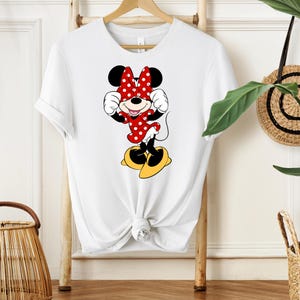 Pu&ograve; includere: T-shirt bianco con un'illustrazione di Minnie Mouse. Minnie indossa un vestito e un fiocco a pois rossi, con scarpe gialle. Il disegno &egrave; centrato sulla maglietta, annodata in vita.