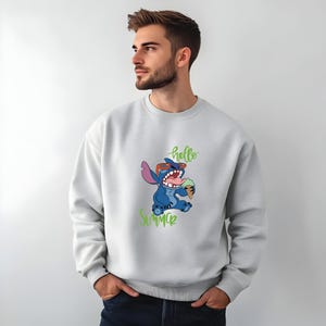 Puede incluir: Sudadera gris claro con un dise&ntilde;o de Stitch sosteniendo un helado. Stitch, un personaje azul, lleva gafas de sol rojas. Las palabras "hello summer" en verde est&aacute;n escritas arriba y abajo.