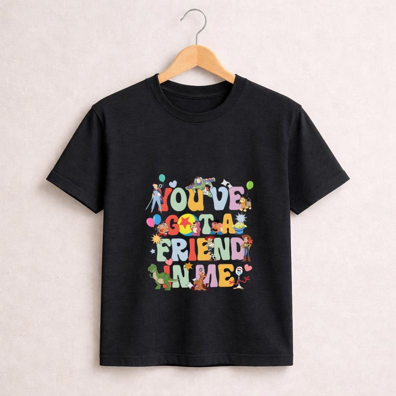May include: Black t-shirt with the phrase "You've Got a Friend in Me" in colorful, playful lettering. The design includes cartoon characters and balloons, creating a fun and whimsical look. The shirt is hanging on a wooden hanger.
