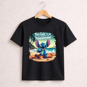 Pu&ograve; includere: T-shirt nera con un personaggio dei cartoni animati su una tavola da surf sulla spiaggia. Il personaggio &egrave; blu con grandi orecchie e sorride. Il testo "Too Cute to be this Troublemaker" &egrave; sopra il personaggio.