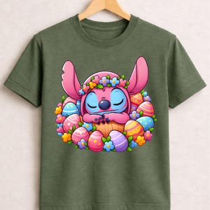 May include: Olive green t-shirt featuring a cartoon character with pink ears and blue eyes, surrounded by colorful Easter eggs and flowers. The character is wearing a flower crown and appears to be sleeping. The design is centered on the shirt.
