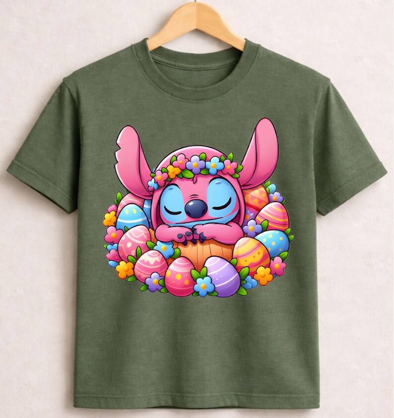 May include: Olive green t-shirt featuring a cartoon character with pink ears and blue eyes, surrounded by colorful Easter eggs and flowers. The character is wearing a flower crown and appears to be sleeping. The design is centered on the shirt.