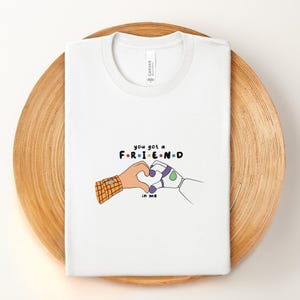 May include: White t-shirt with the text "you got a FRIEND in me" in colorful letters. The design features two cartoon hands forming a heart shape. One hand has a plaid sleeve, the other is a robot hand.