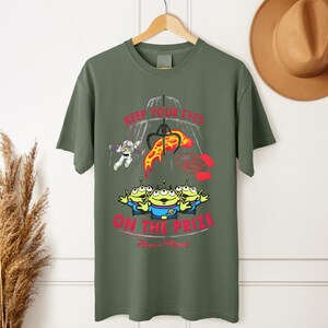 May include: Olive green t-shirt featuring a graphic from the movie Toy Story. The design includes the text "KEEP YOUR EYES ON THE PRIZE" with images of the Little Green Men, Buzz Lightyear, and a claw machine.