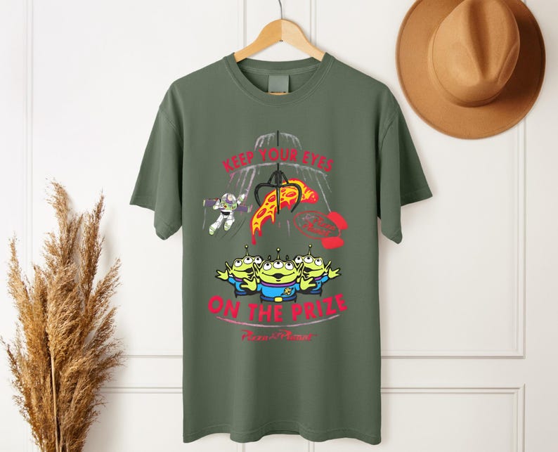 May include: Olive green t-shirt featuring a graphic from the movie Toy Story. The design includes the text "KEEP YOUR EYES ON THE PRIZE" with images of the Little Green Men, Buzz Lightyear, and a claw machine.