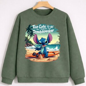 Puede incluir: Sudadera verde oliva con un gr&aacute;fico del personaje Stitch en una tabla de surf en la playa. La imagen incluye el texto "Too Cute to Be This Troublemaker". El dise&ntilde;o est&aacute; ambientado sobre un fondo de palmeras, arena y olas.