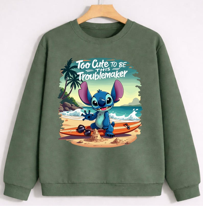 Puede incluir: Sudadera verde oliva con un gr&aacute;fico del personaje Stitch en una tabla de surf en la playa. La imagen incluye el texto "Too Cute to Be This Troublemaker". El dise&ntilde;o est&aacute; ambientado sobre un fondo de palmeras, arena y olas.