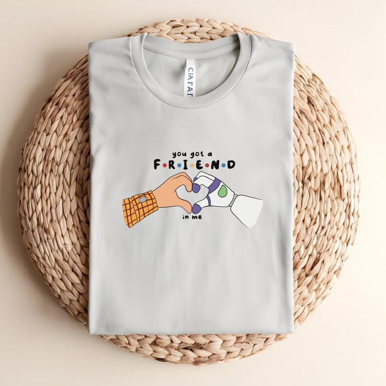May include: Light gray t-shirt featuring a graphic of two hands forming a heart, one human and one robotic, with the text "you got a FRIEND in me" in colorful letters. The shirt is displayed on a woven basket.