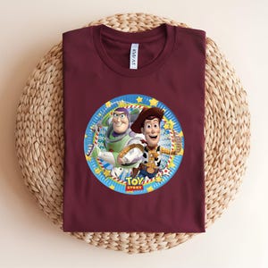 Puede incluir: Una camiseta granate con un gr&aacute;fico circular de Buzz Lightyear y Woody de Toy Story. El gr&aacute;fico est&aacute; enmarcado por un borde azul con estrellas y las palabras "Toy Story". La camiseta se muestra sobre una superficie tejida.