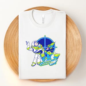 May include: White t-shirt with a graphic of Buzz Lightyear and the Little Green Men from Toy Story. The design includes a blue target, a rocket, and the words "Star Command". The shirt is folded on a wooden surface.