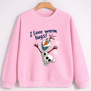 May include: A light pink sweatshirt featuring a cartoon snowman character with the text "I love warm hugs!". The snowman has a carrot nose, twig arms, and a wide smile. Snowflakes are also visible.