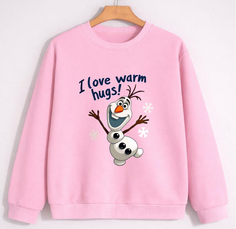 May include: A light pink sweatshirt featuring a cartoon snowman character with the text "I love warm hugs!". The snowman has a carrot nose, twig arms, and a wide smile. Snowflakes are also visible.