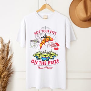 May include: White t-shirt featuring a colorful graphic from the movie Toy Story. The design includes the text "KEEP YOUR EYES ON THE PRIZE" with images of the Pizza Planet aliens, Buzz Lightyear, and a pizza slice.