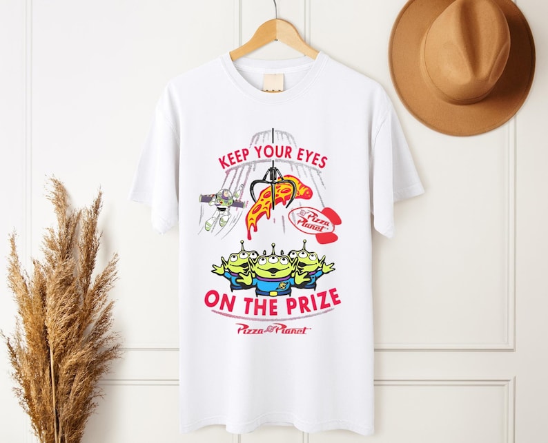May include: White t-shirt featuring a colorful graphic from the movie Toy Story. The design includes the text "KEEP YOUR EYES ON THE PRIZE" with images of the Pizza Planet aliens, Buzz Lightyear, and a pizza slice.