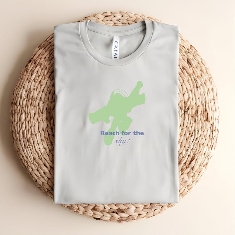 May include: A pale grey t-shirt featuring a green space ranger graphic and the blue text "Reach for the sky!". The shirt is folded on a woven, circular mat.