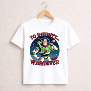 Puede incluir: Camiseta blanca con una ilustraci&oacute;n de dibujos animados de Buzz Lightyear de Toy Story. El dise&ntilde;o muestra a Buzz con su traje espacial, un fondo estrellado y el texto "TO INFINITY... and WHATEVER" en letras rojas y blancas. La camiseta est&aacute; colgada en una percha de madera.