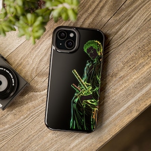 Puede incluir: Funda de teléfono negra con una ilustración verde neón y amarilla de un personaje con pelo verde y espadas. El diseño está sobre un fondo negro. La funda del teléfono está sobre una superficie de madera.