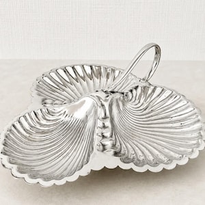 May include: A silver-toned metal serving dish featuring three connected shell-shaped compartments with scalloped edges and a central arched handle. The surface has a polished, reflective finish with embossed radial ridges mimicking the texture of a seashell.