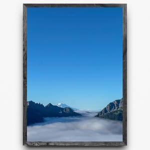 May include: A framed landscape print featuring a mountain range with a snow-capped peak, set against a bright blue sky. The mountains are partially obscured by a layer of fog or clouds, creating a serene and natural scene.