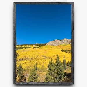 Op de afbeelding: Een ingelijste landschapsfoto met een levendige herfstsc&egrave;ne. De afbeelding toont een bergketen onder een heldere, helderblauwe lucht. De voorgrond is gevuld met gele bladeren, en er zijn groenblijvende bomen verspreid.