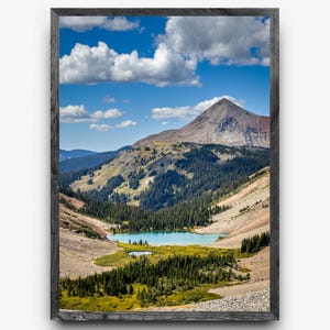 May include: A framed landscape photograph of a mountain range with a lake. The image features a blue sky with white clouds, a mountain peak, and a valley with a lake surrounded by green trees and vegetation.