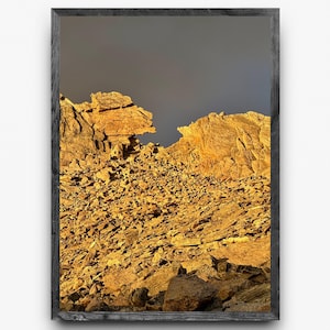 Puede incluir: Una impresión enmarcada que presenta un paisaje de montaña. La imagen muestra una cordillera rocosa de color dorado bajo un cielo oscuro y nublado. La obra de arte está encerrada en un marco oscuro y texturizado, realzando los tonos naturales.