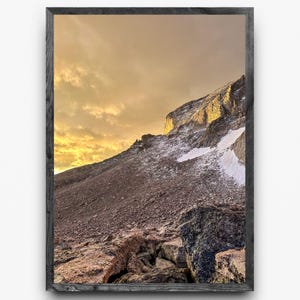 Puede incluir: Fotografía enmarcada de un paisaje de montaña. La imagen captura una ladera rocosa con parches de nieve, iluminada por la luz dorada del sol. El cielo está lleno de nubes de tonos cálidos, creando una vista dramática y pintoresca.