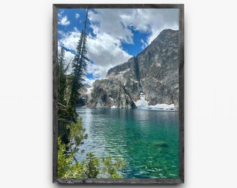 May include: A framed landscape photograph of a mountain lake. The water is a clear turquoise color, reflecting the sky and surrounding mountains. The sky is blue with fluffy white clouds. The mountains are rocky with patches of snow.