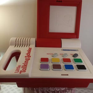 Può includere: Un giocattolo educativo elettronico VTech Little Talking Scholar bianco e rosso con coperchio sollevabile, griglia dell'altoparlante e pannello di controllo con otto pulsanti quadrati colorati, tre pulsanti rettangolari e il nome del prodotto.