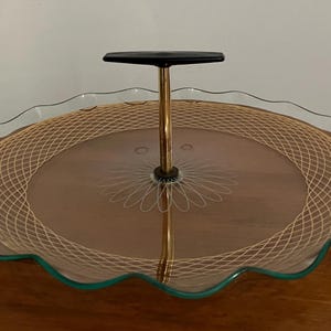 May include: A glass serving tray with a scalloped edge and a gold-coloured lattice design. The tray has a black plastic handle and a gold-coloured metal stand in the centre.