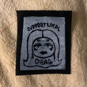 May include: A rectangular patch with a black border. The patch features a stylized illustration of a face with long hair and the words "SUPPORT LOCAL DRAG" in black lettering. The background is light gray.