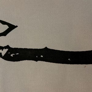 May include: A black ink graphic print of a tactical axe on a light-colored fabric background. The design features a sharp blade, a pointed spike on the opposite side of the head, and a textured handle with visible rivets along the grip.