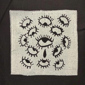 May include: A square fabric patch featuring a light gray background with a repeating pattern of black embroidered eyes. The design includes eleven individual eyes of varying sizes, each with distinct eyelashes and pupils, arranged around a central teardrop shape.