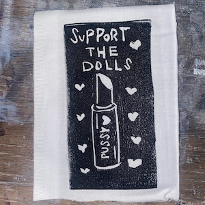 May include: A white cloth with a black print that reads "SUPPORT THE DOLLS" above a lipstick design with the word "PUSSY" on it. White hearts are scattered around the lipstick.
