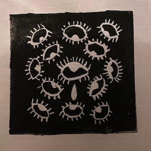May include: A black square print featuring a cluster of white, stylized eyes. The eyes are arranged in a circular pattern, with a single eye at the center and others surrounding it. The print has a handmade, artistic aesthetic.