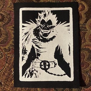 May include: Black and white patch featuring a stylized illustration of a character with spiky hair and a belt with a circular buckle. The character is set against a black background.