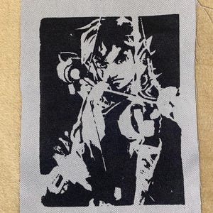 May include: A black and white patch depicting a person holding a weapon. The image is printed on a square, light grey fabric with a textured finish. The artwork has a bold, graphic style, suitable for clothing or accessories.