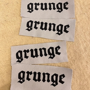 Può includere: Quattro etichette di tessuto grigio con la parola "grunge" stampata in nero con carattere gotico. Le etichette rettangolari sono impilate su una superficie beige. La parola "grunge" è ripetuta su ogni etichetta.