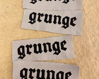 Puede incluir: Cuatro etiquetas de tela gris con la palabra "grunge" impresa en negro con fuente gótica. Las etiquetas rectangulares están apiladas sobre una superficie beige. La palabra "grunge" se repite en cada etiqueta.