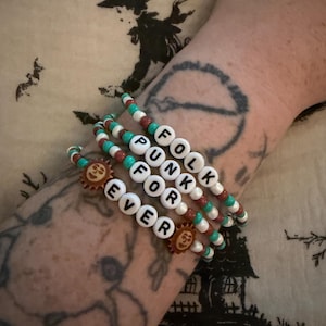 May include: A set of three beaded bracelets featuring white letter beads that spell out FOLK PUNK FOR EVER. The bracelets are constructed with alternating turquoise, brown, and white beads, and include two small brown sun-shaped charms.